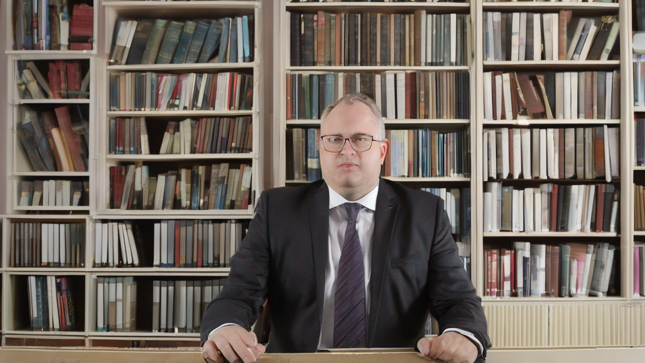 Portret Tomasza Kowalczyka, mężczyzny w garniturze, stojącego przed ścianą książek w bibliotece
